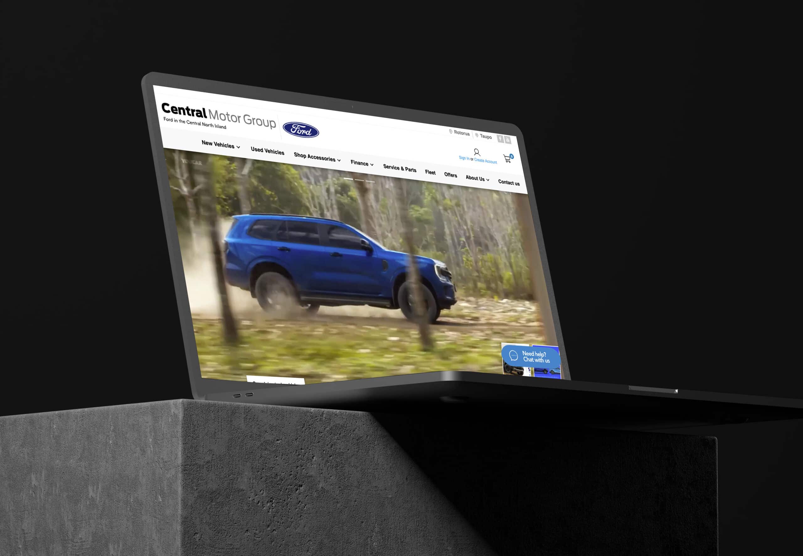 Laptop displaying the Central Motor Group Ford website, featuring a promotional video of a blue Ford SUV driving through a forest trail, with the site’s navigation menu and chat support button visible.