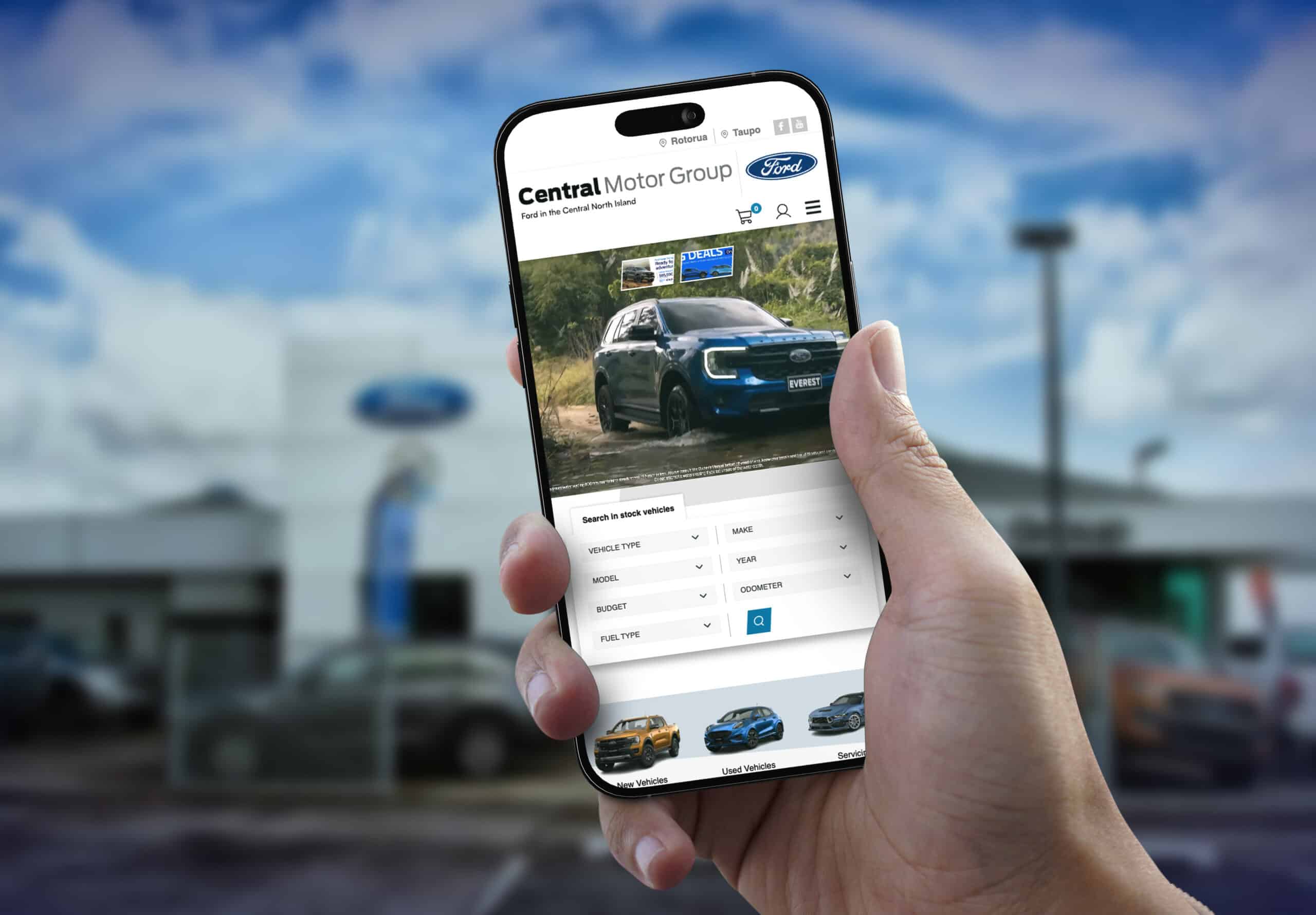Person holding a smartphone displaying the Central Motor Group Ford website, with a blue Ford Everest SUV featured on the homepage, in front of a blurred Ford dealership building.