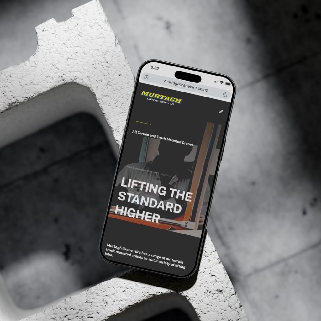 Smartphone displaying the Murtagh Crane Hire website, showing the slogan 'Lifting the Standard Higher' on a dark-themed homepage, placed on a textured concrete block with a dramatic light and shadow contrast.