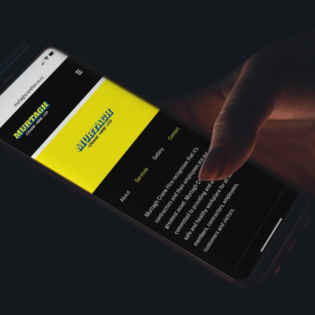 A close-up of a smartphone screen displaying the Murtagh Crane Hire Ltd website in dark mode, with the logo in blue and yellow at the top. The navigation menu lists “About,” “Services,” “Gallery,” and “Contact,” with the Services link highlighted in yellow. Below, white text describes the company’s commitment to providing a safe and healthy workplace for staff, contractors, customers, and visitors. The phone is held in a person’s hand against a dark background.