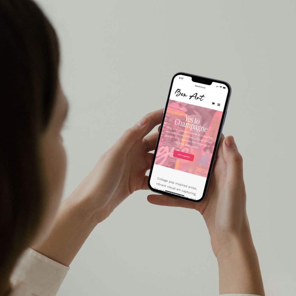 Person holding a smartphone displaying the Bex Art website, featuring a vibrant collage art piece with the text "Yes to Champagne" in white over a pink and orange artistic background.