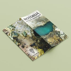 A folded Rotorua Official Visitor Guide brochure lying on a light green surface, featuring an aerial photo of geothermal terrain and a vivid blue hot pool on the cover