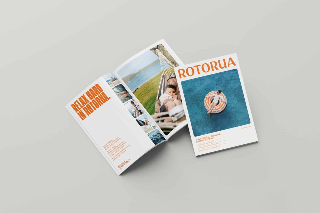 Two travel magazines for Rotorua displayed on a light grey surface. One magazine is closed, showing a cover with bold orange text 'ROTORUA' at the top and a photo of a woman relaxing on a circular float in a blue pool. The other magazine is open, revealing a full-page headline 'RELAX HARD IN ROTORUA' in orange text on the left page and multiple lifestyle images on the right page, including a father and child resting in a hammock near a lake.