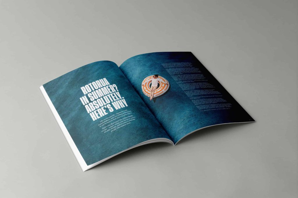 Open magazine on a grey surface featuring a summer-themed Rotorua article. The left page has large white text reading 'Rotorua in Summer? Absolutely. Here’s Why' on a textured blue background. The right page shows a person lounging on an orange-and-white circular float in a pool, with article text below.