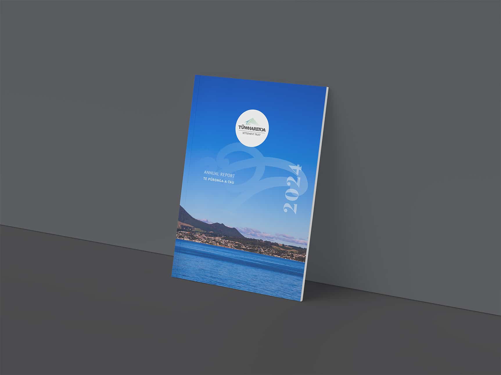 Standing upright against a grey wall, the 2024 Annual Report for Tūwharetoa Māori Trust Board features a vibrant blue cover with the organisation’s white logo at the top, the year “2024” along the side, and a scenic photograph of a lake with a township and mountains in the background.