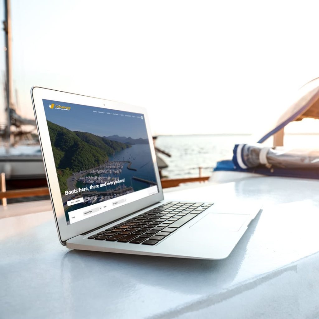 Open silver laptop displaying the Vinings Marine Group website on a boat deck, with the screen showing a marina aerial view and the text "Boats here, there and everywhere!" against a scenic coastal backdrop in bright sunlight.