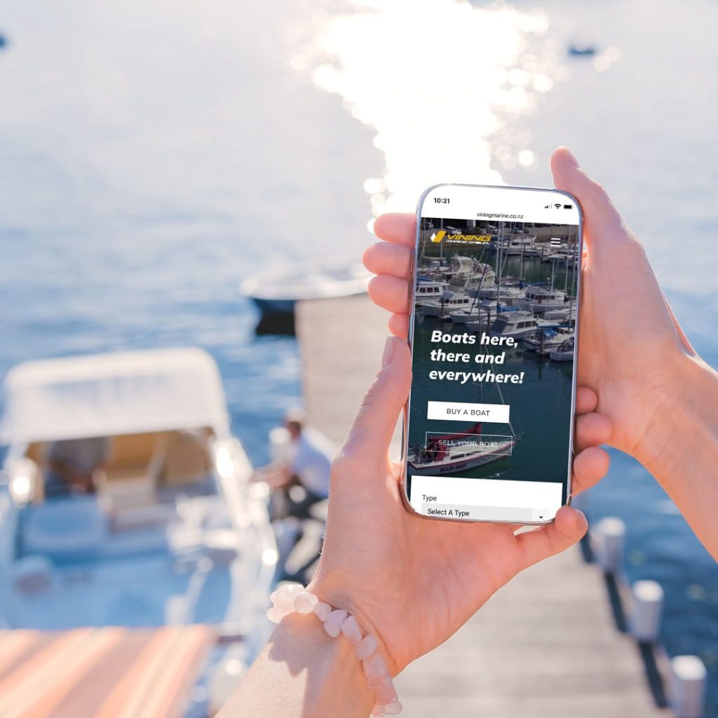 Person holding a smartphone outdoors near a marina, viewing the Vinings Marine Group website homepage, which displays a banner image of boats in a harbor with the headline “Boats here, there and everywhere!” and buttons to buy or sell a boat.