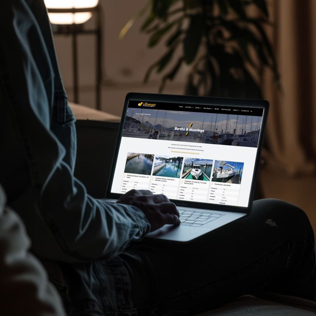 Person using a laptop in a dimly lit room, viewing the Vining Marine Group website’s "Berths & Moorings" page, which displays multiple listings for available marina berths with images, specifications, and prices.